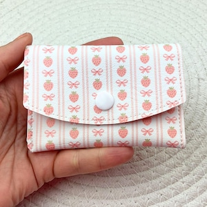 Puede incluir: Una pequeña cartera blanca con cierre de solapa, adornada con un patrón repetido de fresas rosas y lazos sobre un fondo a rayas. La cartera se asegura con un botón blanco.