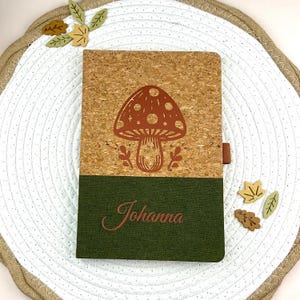 Könnte beinhalten: Ein Notizbuch mit Korkeinband und grünem Unterteil. Der Korkbereich zeigt ein braunes Pilzdesign. Der Name "Johanna" ist in brauner Schrift auf dem grünen Bereich aufgedruckt. Dekorative Blattausschnitte sind um das Notizbuch verstreut.