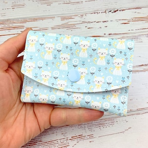 Op de afbeelding: Een lichtblauwe portemonnee met een herhaald patroon van witte katten met gele strikken en bloemen. De portemonnee heeft een klepsluiting met een lichtblauwe knoop. De portemonnee wordt in een hand gehouden.