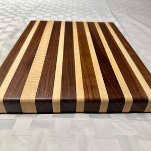 May include: A rectangular wooden cutting board with alternating stripes of light and dark wood. The board has a rounded edge and is made of walnut and maple.