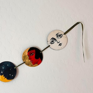 May include: A decorative wall hanging featuring three circular discs connected by a dark green ribbon. The discs display illustrations of a dark starry night, a profile of a person with red skin and black hair, and a pale moon face with detailed eyes.
