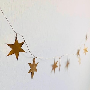 May include: A decorative garland featuring multiple metallic gold-colored star-shaped ornaments suspended from a thin, dark wire. The stars vary in size and are spaced along the length of the wire, creating a hanging arrangement of celestial shapes.