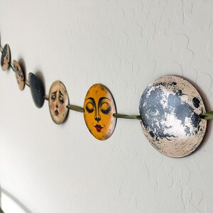 May include: A decorative garland featuring a series of circular discs strung along a green ribbon. Each disc displays a unique illustration of a moon face with varying expressions, colors, and textures, ranging from golden yellow to dark grey.