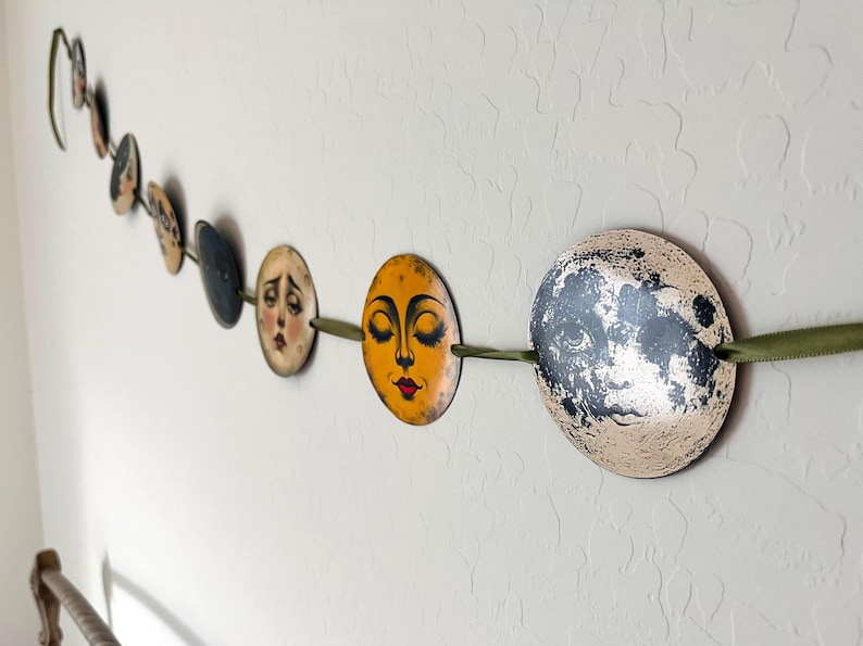 May include: A decorative garland featuring a series of circular discs strung along a green ribbon. Each disc displays a unique illustration of a moon face with varying expressions, colors, and textures, ranging from golden yellow to dark grey.