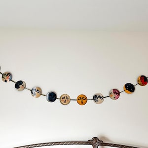 May include: A decorative garland featuring eleven circular discs strung together on a dark ribbon. Each disc displays a unique illustration of a face or celestial body, including various moon phases, sun motifs, and stylized portraits in diverse colors.