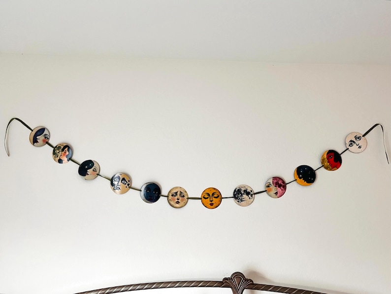 May include: A decorative garland featuring eleven circular discs strung together on a dark ribbon. Each disc displays a unique illustration of a face or celestial body, including various moon phases, sun motifs, and stylized portraits in diverse colors.