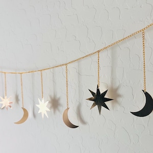 May include: A gold-colored metal garland featuring a horizontal chain with dangling celestial charms. The charms include various sizes of crescent moons and multi-pointed stars, all suspended from the main chain by individual short gold chains.