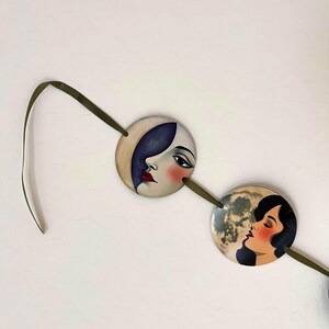 May include: Two circular ornaments connected by a dark olive green ribbon. The left ornament features a stylized face with a dark blue crescent moon shape, while the right ornament depicts a profile of a person against a full moon background.