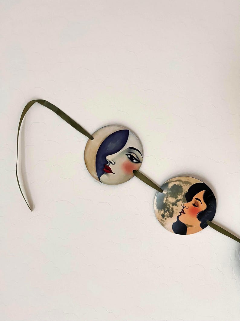 May include: Two circular ornaments connected by a dark olive green ribbon. The left ornament features a stylized face with a dark blue crescent moon shape, while the right ornament depicts a profile of a person against a full moon background.