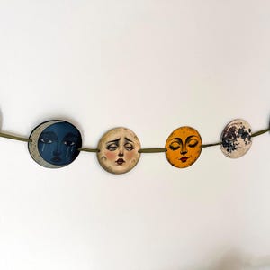 May include: A decorative garland featuring a series of circular discs suspended on a green cord. Each disc displays a unique illustration of a moon with a human face, rendered in various colors including beige, dark blue, yellow, and white.
