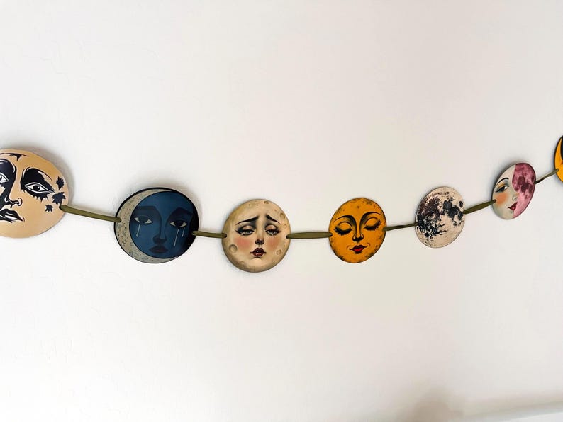 May include: A decorative garland featuring a series of circular discs suspended on a green cord. Each disc displays a unique illustration of a moon with a human face, rendered in various colors including beige, dark blue, yellow, and white.