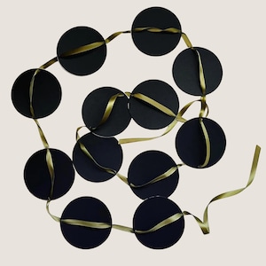 May include: A decorative garland consisting of eleven solid black circular discs strung together on a continuous olive green satin ribbon. The ribbon weaves through each circular piece, creating a connected chain of black circles and green ribbon.