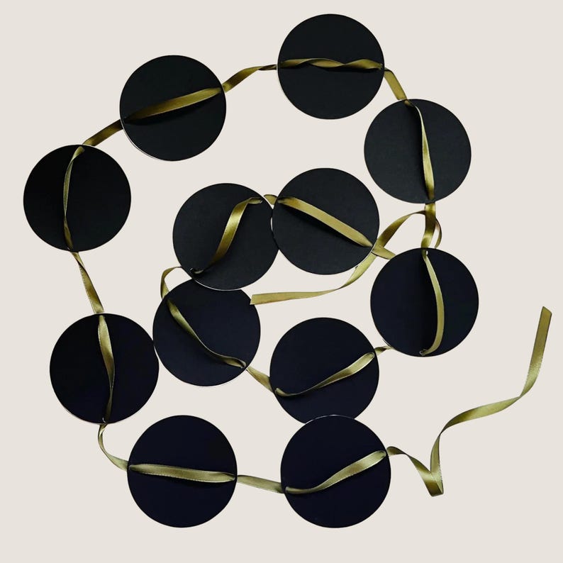 May include: A decorative garland consisting of eleven solid black circular discs strung together on a continuous olive green satin ribbon. The ribbon weaves through each circular piece, creating a connected chain of black circles and green ribbon.