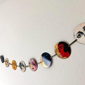 May include: A decorative garland featuring a series of circular discs strung together on a green ribbon. Each disc displays a unique illustration, including stylized faces with expressive features, celestial motifs, and vibrant colors like red and gold.