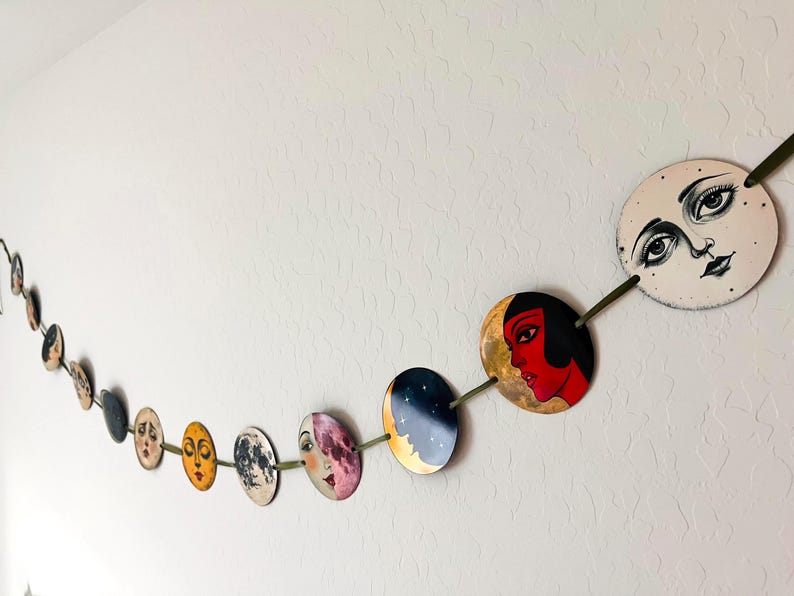 May include: A decorative garland featuring a series of circular discs strung together on a green ribbon. Each disc displays a unique illustration, including stylized faces with expressive features, celestial motifs, and vibrant colors like red and gold.