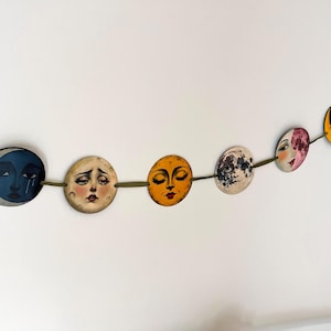 May include: A decorative garland featuring a series of circular discs connected by a green ribbon. Each disc displays a unique illustration of a moon with a human face, rendered in various colors including blue, cream, yellow, and textured grey tones.