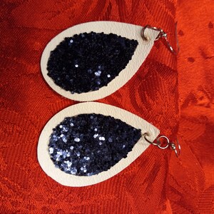 Può includere: Un paio di orecchini a forma di goccia con base in pelle color crema e dettagli in glitter nero.