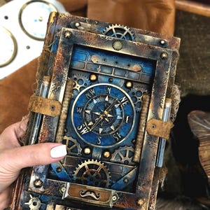 May include: A steampunk journal with a blue and bronze colour scheme. The cover features a compass design, gears, and a decorative moustache. The journal is bound with leather straps and has a vintage, aged appearance.