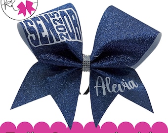 May include: A dark blue glitter hair bow featuring the text SENIOR 2025 in white block letters on one side and the name Alevia in white script on the other. The center of the bow is embellished with a vertical band of small, clear rhinestones.
