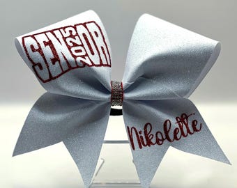 May include: White cheer bow with red glitter lettering that says "Senior 2023" and "Nikolete".