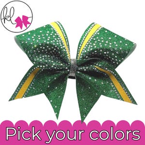 May include: A large hair bow featuring green glitter fabric with a yellow stripe border and scattered clear rhinestones. The center of the bow is wrapped in a band of silver rhinestones. The text "Pick your colors" appears at the bottom.