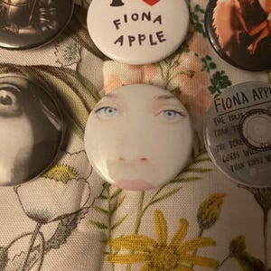 May include: A collection of round buttons featuring images and text. One button displays a face with blue eyes and pink lips. Other buttons have the text "FIONA APPLE" and album art. The buttons are on a floral patterned fabric.