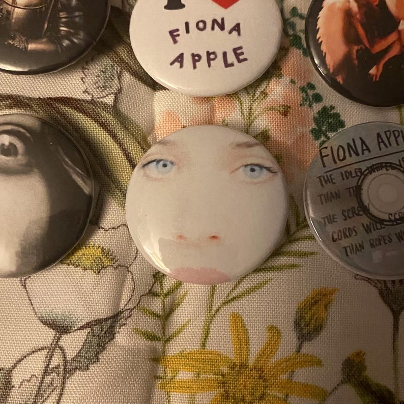 May include: A collection of round buttons featuring images and text. One button displays a face with blue eyes and pink lips. Other buttons have the text "FIONA APPLE" and album art. The buttons are on a floral patterned fabric.