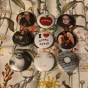May include: A collection of round buttons featuring images and text. Some buttons display portraits, while others have text like "FIONA APPLE" and "I ❤️ FIONA APPLE". One button shows a red apple with the name "FIONA".