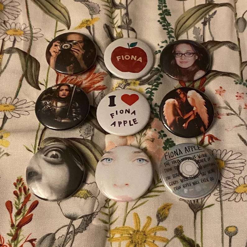 May include: A collection of round buttons featuring images and text. Some buttons display portraits, while others have text like "FIONA APPLE" and "I ❤️ FIONA APPLE". One button shows a red apple with the name "FIONA".