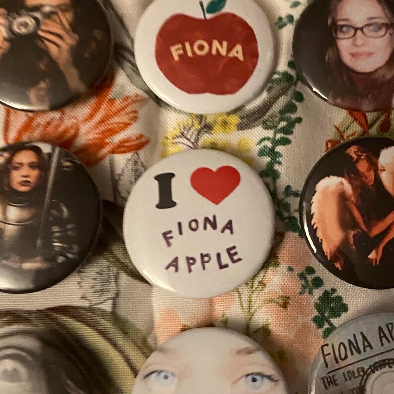 May include: A collection of round buttons featuring images and text. One button has a red apple with the name "FIONA" in white. Another button says "I ❤️ FIONA APPLE". Other buttons have images of a person.