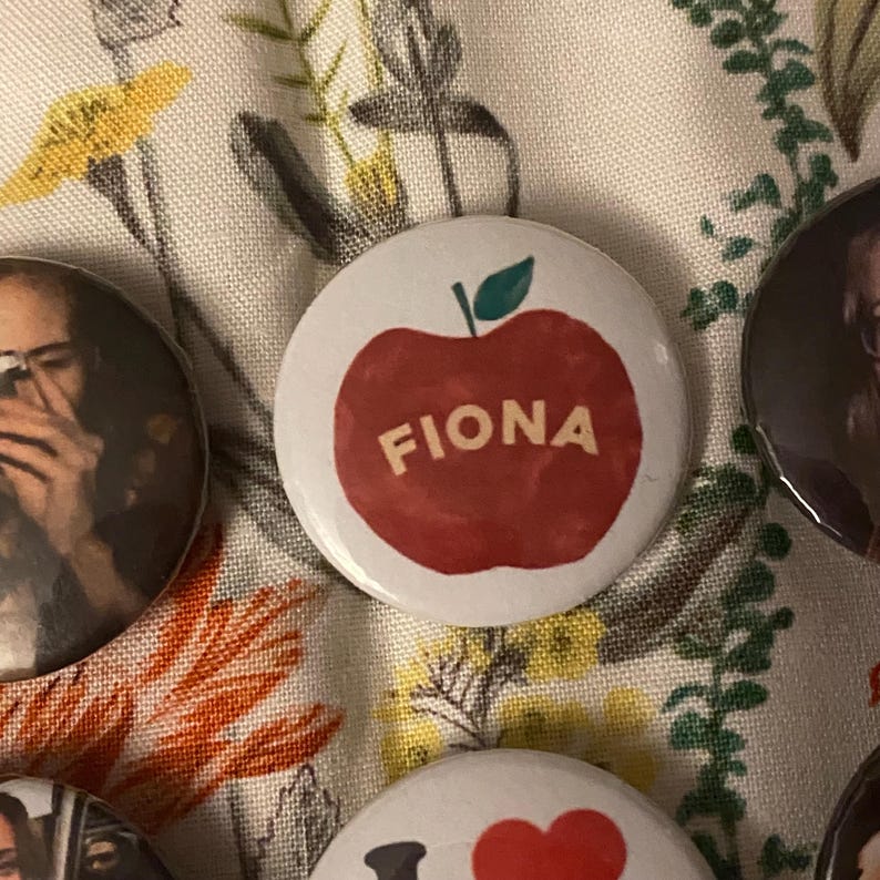 May include: A collection of round buttons with various designs. One button features a red apple with the name "FIONA" in a serif font. Other buttons have images and text.