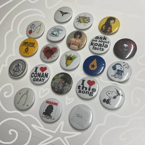 May include: A collection of round pinback buttons with various designs and text. Some buttons feature illustrations, such as a bat, Snoopy, and a wishbone. Other buttons have text like "Employee of the Month," "I love this song," and "Missing Alley Rose."