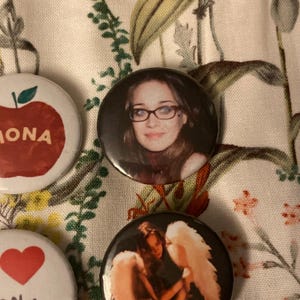 May include: A collection of round pinback buttons with various designs. One button features a woman wearing glasses, another has an apple with the name "MONA", and a third displays a heart. The buttons are on a floral patterned fabric.