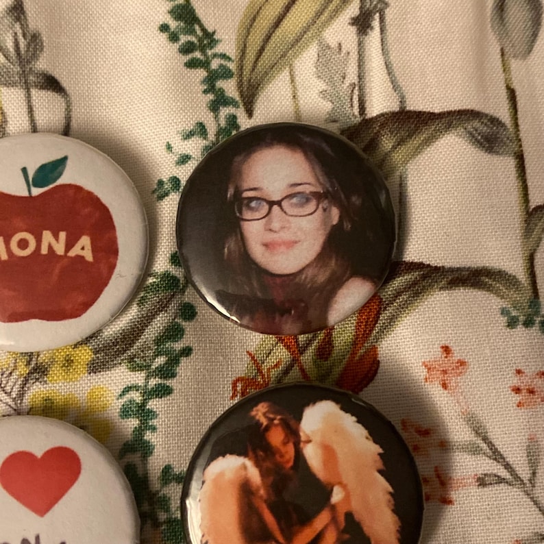 May include: A collection of round pinback buttons with various designs. One button features a woman wearing glasses, another has an apple with the name "MONA", and a third displays a heart. The buttons are on a floral patterned fabric.