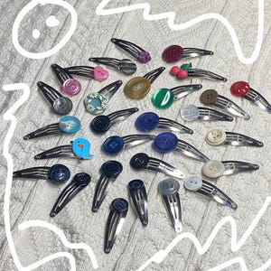 May include: A collection of silver metal hair barrettes, each adorned with a unique decorative button. Buttons vary in color and design, including floral patterns, hearts, and solid colors. The barrettes are arranged on a textured, off-white surface.