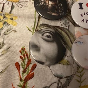 May include: A collection of round pinback buttons with various designs. One button features a black and white close-up of an eye. Other buttons display images and text, including an image of a figure and the text "I LOVE FLOWERS".