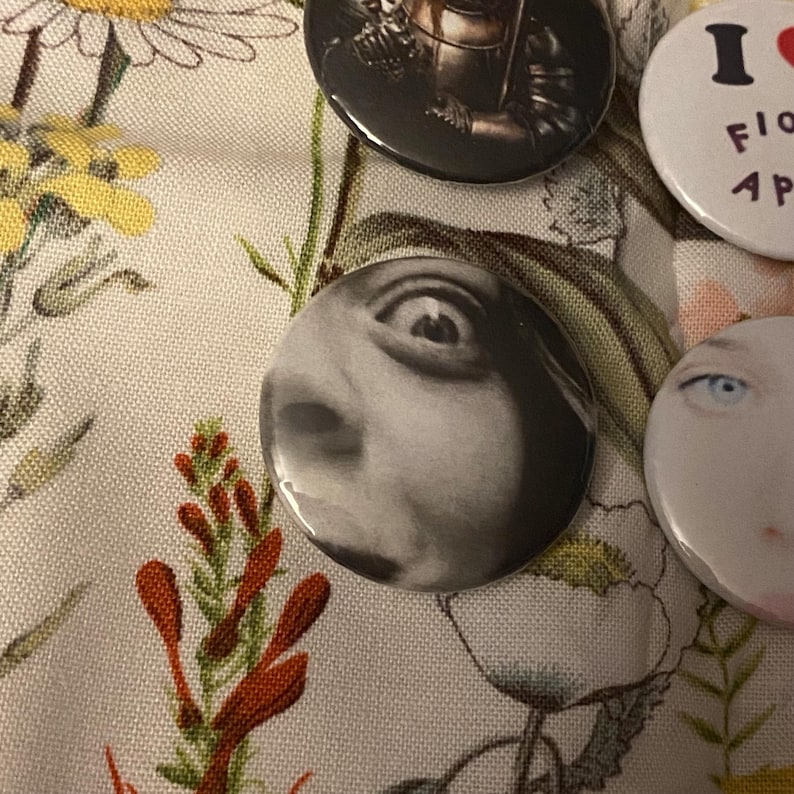 May include: A collection of round pinback buttons with various designs. One button features a black and white close-up of an eye. Other buttons display images and text, including an image of a figure and the text "I LOVE FLOWERS".