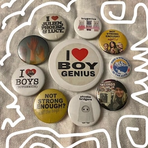 May include: A collection of round buttons with various designs and text. Some buttons feature the text "I BOY GENIUS", "I BOYS (BOYGENIUS)", and "NOT STRONG ENOUGH? call the boys!". Other buttons have images and text related to music.