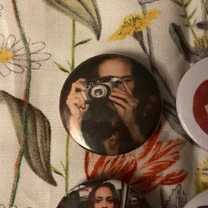 May include: A round button featuring a person holding a vintage camera. The button has a glossy finish and a dark color palette. Other buttons are visible in the image, with a floral background.