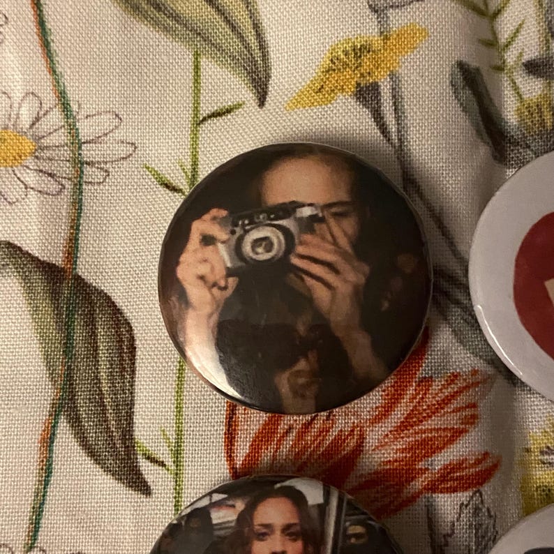May include: A round button featuring a person holding a vintage camera. The button has a glossy finish and a dark color palette. Other buttons are visible in the image, with a floral background.