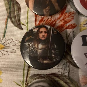 May include: A collection of round buttons with images. One button features a woman in medieval armor holding a sword. The buttons are on a floral patterned fabric.