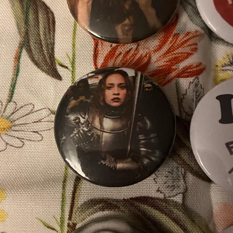 May include: A collection of round buttons with images. One button features a woman in medieval armor holding a sword. The buttons are on a floral patterned fabric.