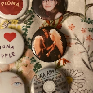 May include: A collection of round buttons featuring images and text. One button has a red apple and the name "FIONA" in white. Another button has a heart and the words "IONA APPLE". Other buttons have images of a woman and text.