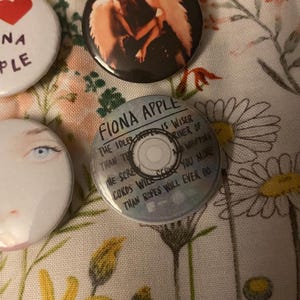 May include: A collection of round buttons with various designs. One button features the text "FIONA APPLE" and song lyrics. Other buttons display images of a face, a heart, and an image of two people. The buttons are on a floral patterned fabric.