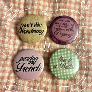 May include: Four circular pin-back buttons with text. Top left is tan with 'Don't die Wondering'. Top right is purple with 'are you only hanging out with me because of the Prophecy?'. Bottom left is light pink with 'pardon my French'. Bottom right is green gingham with 'this is a Button'.