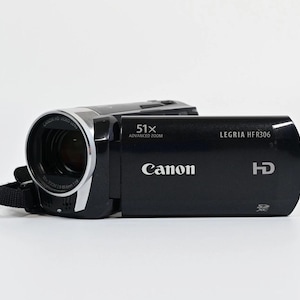 Pode incluir: Uma câmara de vídeo Canon Legria HF R306 preta com lente de zoom avançado de 51x, capacidades de gravação de vídeo HD e ranhura para cartão de memória SDXC. O dispositivo tem um corpo compacto com detalhes prateados e marca em branco.
