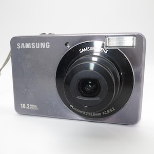 May include: A grey Samsung digital camera featuring a 10.2 megapixels label, a black lens housing with 3x zoom 6.2-18.6mm 1:2.8-5.2 text, a built-in flash, and a light-colored wrist strap attached to the side of the rectangular camera body.