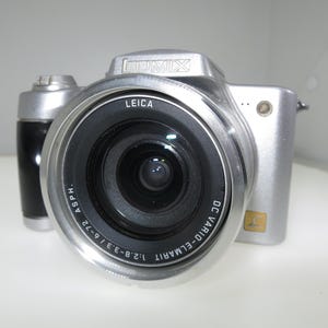 May include: Silver digital camera with a black lens and a black grip. The camera has the word "LEICA" printed on the lens and "LUMIX" on the top. The lens has the following printed on it: "DC VARIO-ELMARIT" and "1:2.8-3.3/6-72".