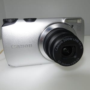 May include: Silver Canon digital camera with a black lens. The camera has a 5x zoom lens and a built-in flash. The Canon logo is visible on the front of the camera. The lens is 50-25.0mm.