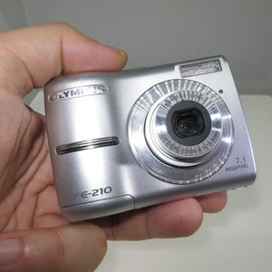 Puede incluir: Cámara digital plateada Olympus FE-210 con una resolución de 7,1 megapíxeles. La cámara tiene un flash incorporado y un objetivo con zoom óptico. La cámara se sostiene en una mano.